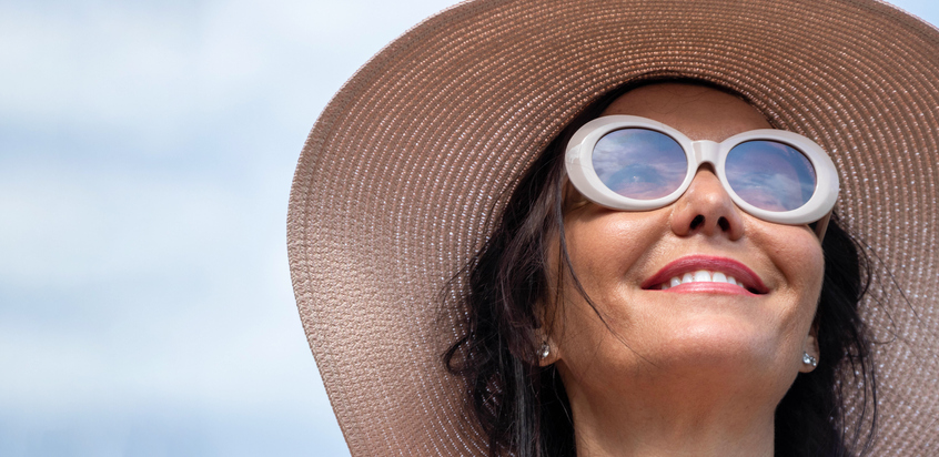 Woman with hat and sunglasses, smilling, on a summer day, with blue sky in the background.